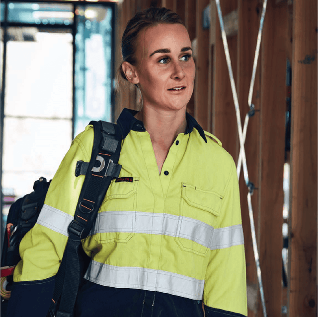 **Alt Text:** Female tradie wearing hi-vis yellow and navy work shirt with reflective tape – custom printed safety shirt with free logo included, compliant with AS/NZS standards.