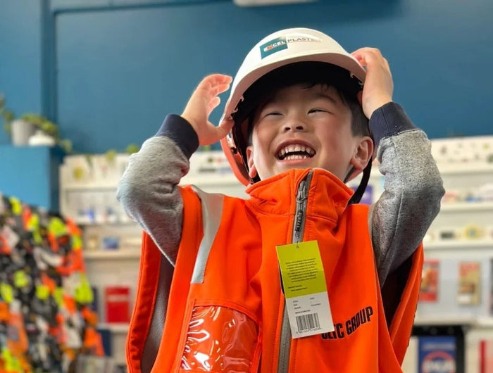 A smiling child wearing an oversized high-visibility orange and navy work vest and white hard hat inside the My Promo Shop showroom. Showcasing the fun side of custom workwear, this playful moment highlights My Uniform Shop’s expertise in high-quality branded safety wear, with over 20 years of experience serving Australian businesses from our Huntingdale, Victoria showroom.