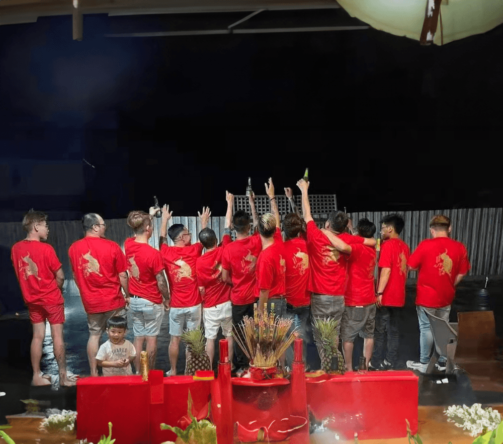 Group of people wearing matching red T-shirts with a gold dragon design, standing in a row with raised hands during a night-time celebration, with a red altar setup in the foreground.