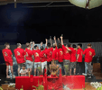 Group of people wearing matching red T-shirts with a gold dragon design, standing in a row with raised hands during a night-time celebration, with a red altar setup in the foreground.
