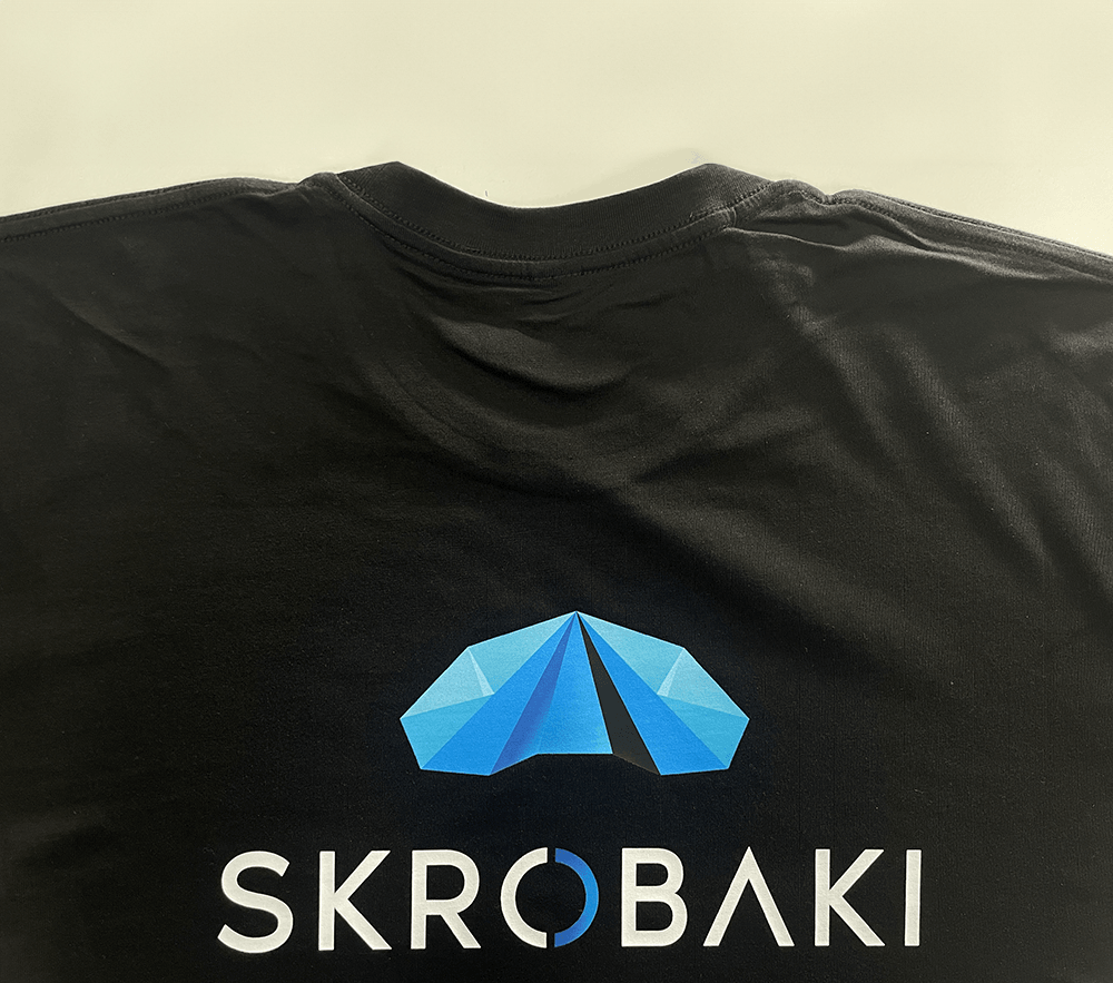 Printed logo of SKROBAKI on the back of a black T-shirt, featuring a geometric blue tent-like graphic above the brand name.