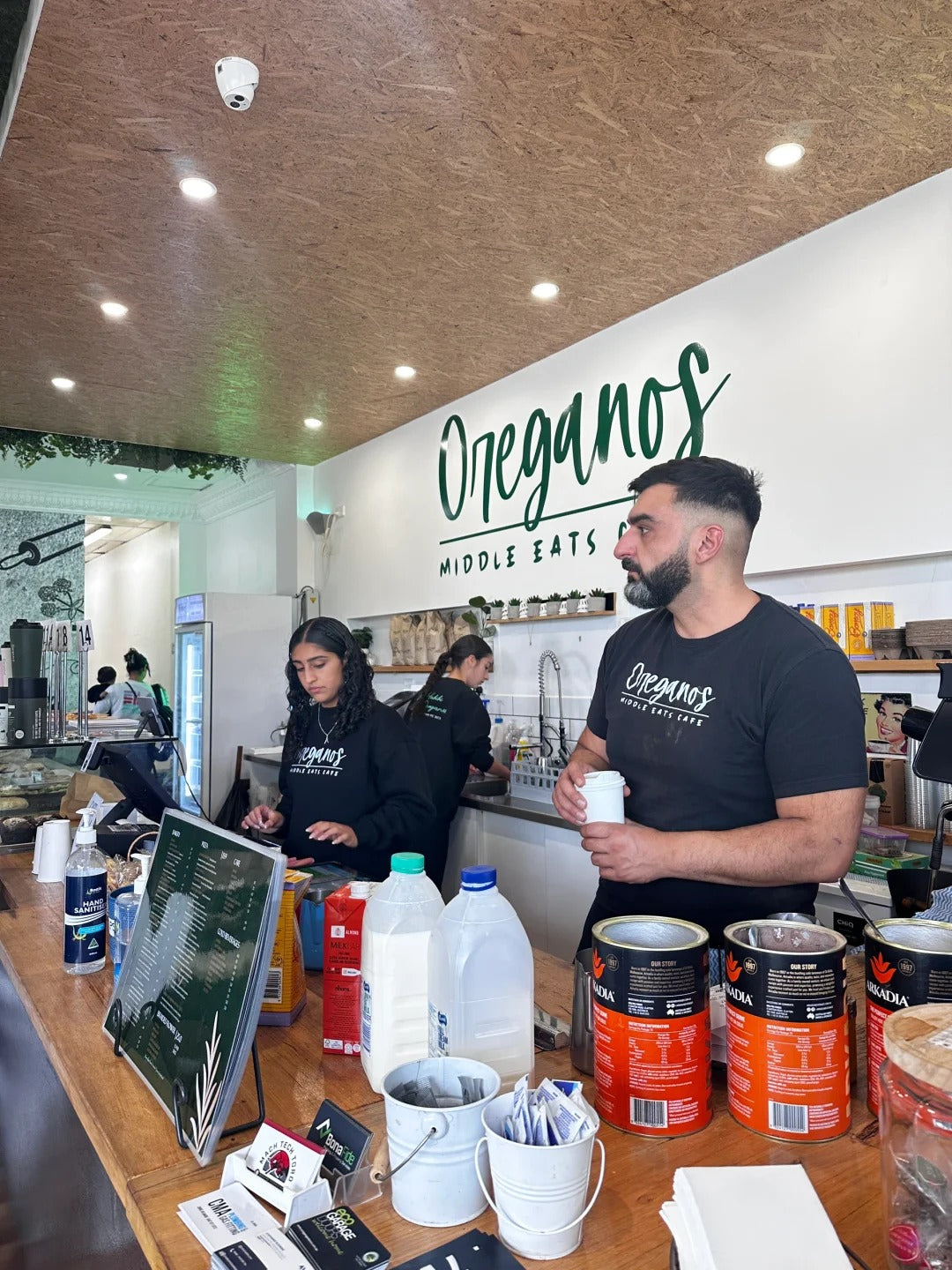 Staff at Oreganos Middle Eats Café wearing custom black cotton uniforms with minimalist logo, behind the counter preparing drinks in a modern café setting.