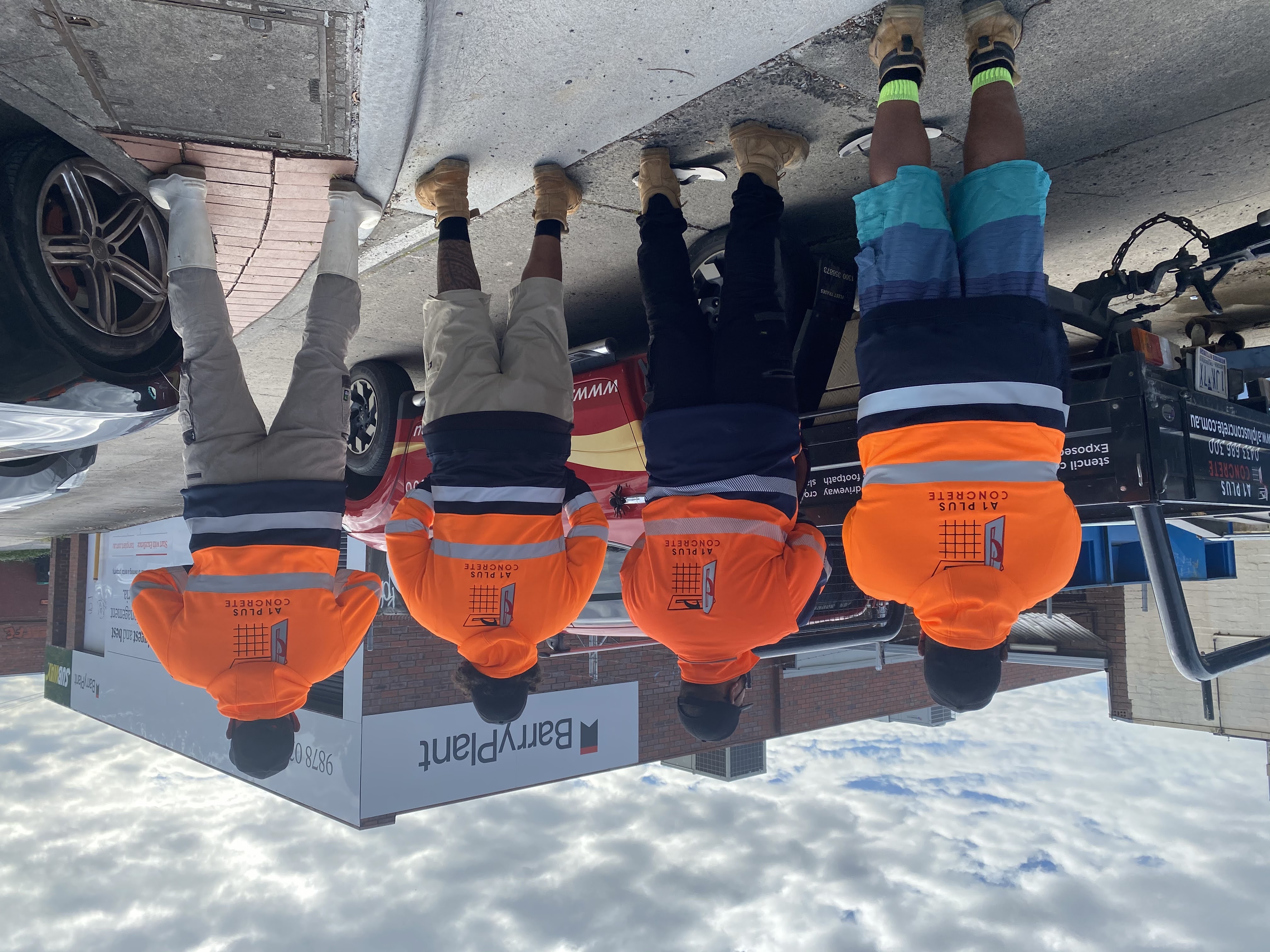 Group of workers wearing custom high-vis orange hoodies with the A1 Plus Concrete logo printed on the back, standing in front of a Barry Plant building.