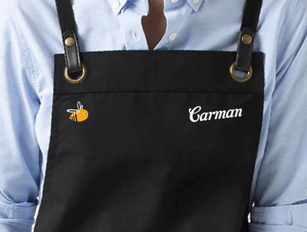 Close-up of a black apron featuring white embroidered name “Carman” and a cute orange bee logo, worn over a light blue shirt. A personalised design by My Uniform Shop, showcasing custom embroidery for hospitality uniforms and staff apparel.