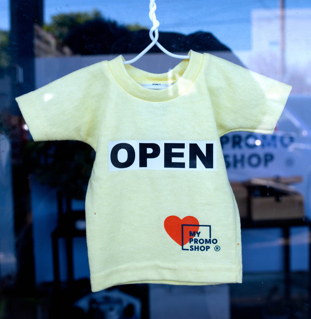 A small yellow T-shirt with a bold “OPEN” print and the My Promo Shop logo, displayed on a hanger in the shop window. A creative and welcoming sign showcasing the fun and custom branding options available at My Promo Shop.